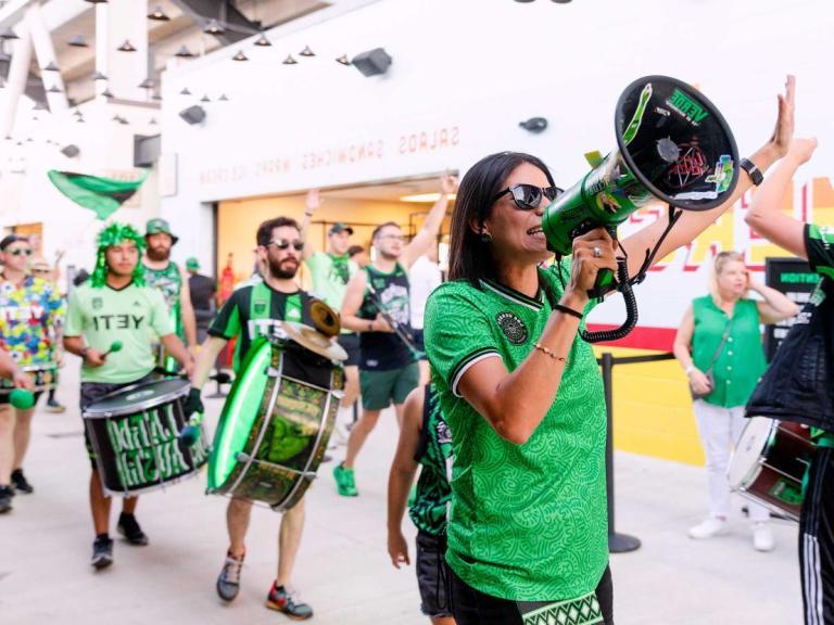 The image shows a group of enthusiastic fans, wearing green and black attire with "YETI" logos, celebrating and marching with drums and a megaphone inside a stadium. The scene is lively, with one person leading chants through the megaphone while others play drums and high-five. The overall atmosphere is energetic and festive, indicating a sports event or rally.