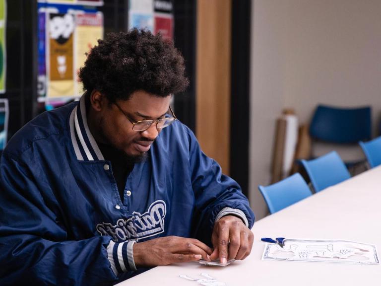 The image shows a man with glasses and a beard, wearing a blue "Patriots" jacket, sitting at a table and concentrating on a craft project involving paper cutouts. He is working alone in a room with blue chairs and colorful posters on the walls, indicating a creative or educational setting.